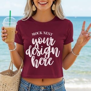 Può includere: Un crop top bordeaux con la scritta "MOCK NEST your design here" in corsivo bianco. Il top è abbinato a jeans blu. L'immagine include anche una bevanda in un bicchiere di plastica trasparente con una cannuccia verde e una borsa tote intrecciata.