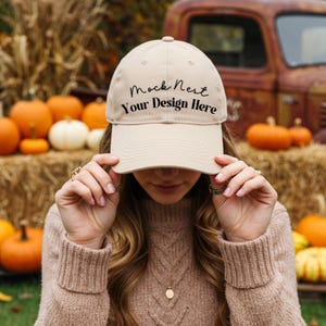 May include: A beige baseball cap with the text "Mock Nest Your Design Here" in black script. The cap is being held up by a person. Pumpkins and hay bales are in the background.