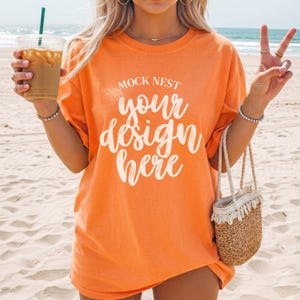 May include: Orange t-shirt with the text "MOCK NEST your design here" in white script. The shirt is worn on a beach. A small straw bag with a white lace trim is held in the hand. A clear plastic cup with a green straw is held in the other hand.