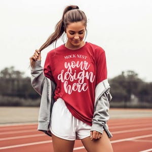 May include: A red long-sleeve shirt with the text "MOCK NEST your design here" in white script. The shirt is paired with white shorts and a gray zip-up jacket. The image is taken on a running track.