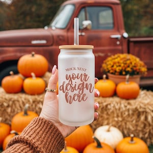 Pode incluir: Um copo de vidro fosco em forma de lata com tampa de bambu e canudo transparente. O copo tem o texto "MOCK NEST your design here" na cor cobre. O fundo inclui abóboras, fardos de feno e uma caminhonete vermelha vintage.