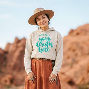 Op de afbeelding: Een persoon draagt een beige hoodie met de tekst "MOCK NEST your design here" in turquoise. De persoon draagt ook een bruine hoed, oorbellen, ringen en een roestkleurige rok. De achtergrond toont een woestijnlandschap.
