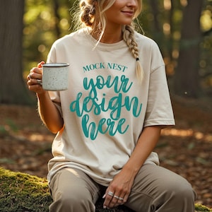 May include: A beige t-shirt with the text "MOCK NEST your design here" in teal. The person is holding a speckled white mug with a black rim. The person is wearing brown pants and sitting on a mossy log.