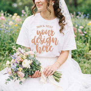 Peut inclure: Un t-shirt blanc avec le texte "MOCK NEST your design here" en couleur cuivre. La personne tient un bouquet de fleurs avec des fleurs blanches, roses et bleues. La personne porte une jupe en tulle blanche et un voile.