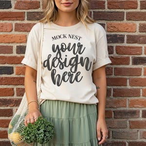May include: A cream-colored t-shirt with rolled-up sleeves featuring the text "MOCK NEST your design here" in black script. The person is wearing a green skirt and holding a mesh bag with produce in front of a brick wall.