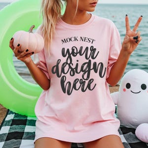 Op de afbeelding: Lichtroze T-shirt met de zwarte tekst "MOCK NEST your design here". De afbeelding bevat ook een lichtroze pompoen, een groene opblaasbare ring en een wit spookvormig pluche speelgoed.