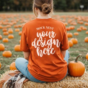 Puede incluir: Una camiseta naranja con el texto "MOCK NEST your design here" en cursiva. La persona está sentada en una paca de heno en un campo de calabazas. La camiseta es de color naranja liso.