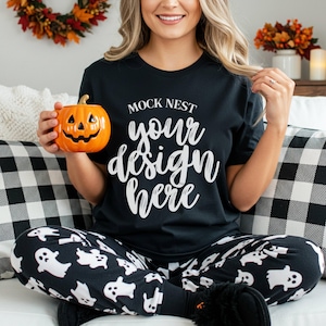 以下が含まれることがあります： 「MOCK NEST your design here」の文字が入った黒いTシャツ。黒と白のゴースト柄のパジャマパンツを履き、オレンジ色のカボチャ型の陶器容器を持っています。背景には白黒のチェック柄のクッションがあります。