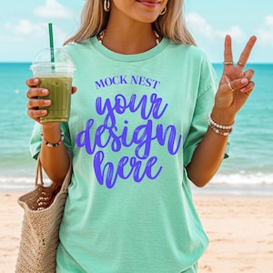 Könnte beinhalten: Ein hellgrünes T-Shirt mit dem Text "MOCK NEST your design here" in Blau. Die Person hält ein grünes Getränk in einem durchsichtigen Plastikbecher mit einem grünen Strohhalm und einer gewebten Tragetasche. Der Hintergrund ist ein Strand mit blauem Wasser.