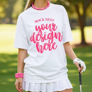 以下が含まれることがあります： ピンク色の「MOCK NEST your design here」の文字が入った白いTシャツ。白いプリーツスカートとピンクのビーズのジュエリーがコーディネートされています。白い手袋をはめた手でゴルフクラブを持っています。背景は緑のゴルフコースです。