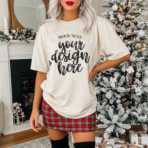 Puede incluir: Camiseta beige con el texto "MOCK NEST your design here" en escritura negra. La modelo lleva una falda a cuadros rojos y verdes y botas negras. Un árbol de Navidad está en el fondo.
