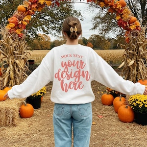 May include: A white sweatshirt with the text "MOCK NEST your design here" in a pink script. The person is standing in a fall-themed setting with pumpkins, hay bales, and an archway of autumn foliage. The person is wearing blue jeans.