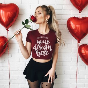Può includere: Un crop top bordeaux con la scritta "MOCK NEST your design here" è indossato con una gonna nera e collant a cuore. Una rosa rossa è tenuta vicino al viso, con palloncini rossi a forma di cuore sullo sfondo.