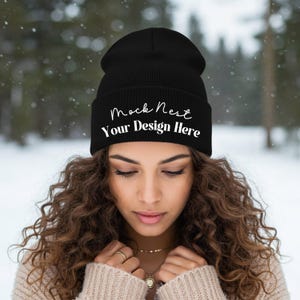 May include: Black beanie with the text "Mock Nest Your Design Here" in white script. The hat is worn by a person with curly brown hair, with a snowy forest background.