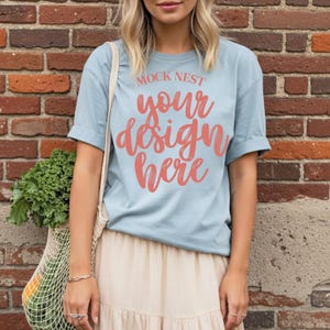 May include: A light blue t-shirt with rolled-up sleeves featuring the text "MOCK NEST your design here" in coral script. The person is wearing a beige skirt and carrying a reusable shopping bag with produce.