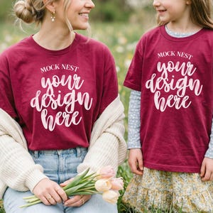Può includere: Due persone indossano magliette bordeaux abbinate con la scritta bianca "MOCK NEST your design here". Una persona tiene in mano un mazzo di fiori rosa e gialli. L'altra persona indossa una camicia a maniche lunghe sotto.