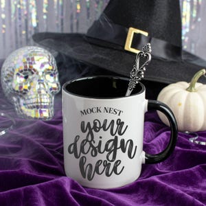 May include: A white coffee mug with a black interior and handle, featuring the text "MOCK NEST your design here." A decorative spoon rests inside the mug. A black witch's hat and a small white pumpkin are in the background, along with a mirrored skull.