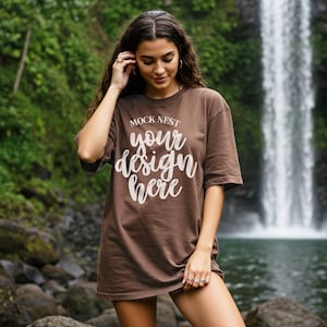 Könnte beinhalten: Ein braunes T-Shirt mit dem Text "MOCK NEST your design here" in weißer Schrift. Das Model steht vor einem Wasserfall. Das Shirt ist einfarbig und hat kurze Ärmel.