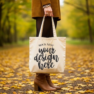 Może przedstawiać: Kremowa torba typu tote z napisem "MOCK NEST your design here" w czarnym skrypcie. Torba jest trzymana przez osobę ubraną w brązowy sweter i buty, stojącą na ścieżce pokrytej żółtymi liśćmi.
