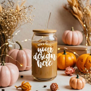 Op de afbeelding: Een heldere glazen beker met een bamboe deksel en een glazen rietje, gevuld met een koffiekleurige drank. De tekst "MOCK NEST your design here" staat op de voorkant. De beker is omgeven door roze en oranje pompoenen en herfstdecoraties.