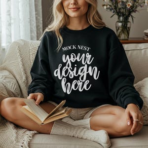 Puede incluir: Sudadera negra de cuello redondo con el texto "MOCK NEST your design here" en escritura blanca. La sudadera la lleva una persona sentada en un sofá beige y sosteniendo un libro.