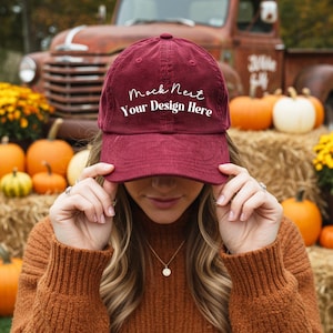 Könnte beinhalten: Burgunderfarbene Cord-Baseballkappe mit dem weißen Text "Mock Nest Your Design Here". Die Kappe wird von einer Person gehalten. Kürbisse und Heuballen befinden sich im Hintergrund.