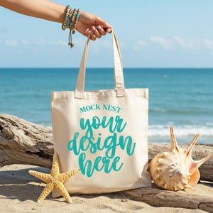 May include: A beige tote bag with the text "MOCK NEST your design here" in teal script. The bag is held by a person's hand, with a beach scene in the background. A starfish and seashell are next to the bag on the sand.
