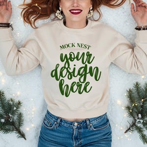 Puede incluir: Sudadera beige de cuello redondo con el texto verde "MOCK NEST your design here". La modelo lleva vaqueros azules. El fondo incluye ramas de abeto y luces de hadas.