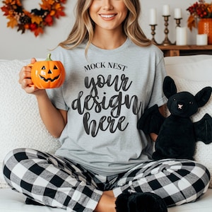 May include: A light gray t-shirt with the text "MOCK NEST your design here". The person is wearing black and white plaid pajama pants and holding an orange pumpkin mug and a black bat plush toy.