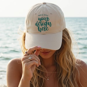 May include: A beige baseball cap with the text "MOCK NEST your design here" in teal. The cap is worn by a person with long blonde hair, with the ocean in the background.