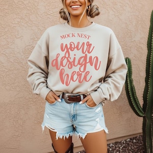 May include: A beige crewneck sweatshirt with the text "MOCK NEST your design here" in pink script. The sweatshirt is paired with light blue denim shorts and a brown belt with a silver buckle. A cactus is visible in the background.