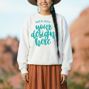 以下が含まれることがあります： 白のスウェットシャツ。 ターコイズ色のスクリプトで「MOCK NEST your design here」と書かれています。 モデルは茶色の帽子と長い茶色のスカートを着用しています。 背景はぼやけた屋外のシーンです。
