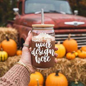 May include: A clear glass tumbler with a bamboo lid and a silver straw, filled with a chocolate beverage and topped with whipped cream. The text "your design here" is displayed on the glass. The background includes pumpkins and a red truck.