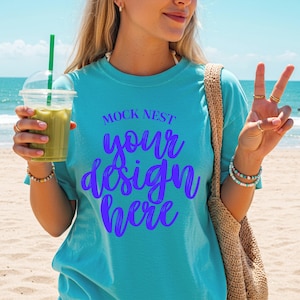 Puede incluir: Una camiseta turquesa con el texto "MOCK NEST your design here" en escritura morada. La persona sostiene un vaso de plástico transparente con una bebida verde y una pajita. La persona lleva pulseras y anillos.