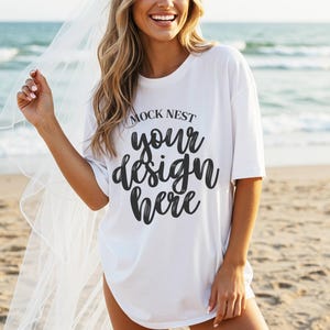 Può includere: T-shirt bianca con la scritta "MOCK NEST your design here" in nero. La t-shirt è indossata su una spiaggia con l'oceano sullo sfondo. La modella sorride e indossa un velo.