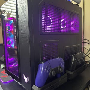 May include: A black gaming computer tower with a glass side panel and purple interior lighting. A purple and a black game controller are docked on the front. The tower has the text "TUF GAMING" on the side.