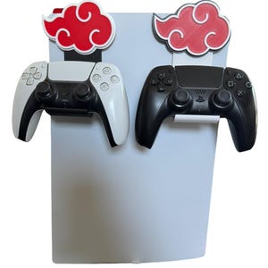 May include: A white and black PlayStation 5 controller and a black controller are displayed on a light gray stand. Each controller is held by a white bracket and topped with a red cloud design.