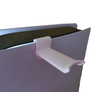May include: A white plastic accessory designed to attach to the edge of a device. The accessory has a curved, handle-like extension. The device appears to be a gaming console, with a white exterior and a dark interior.