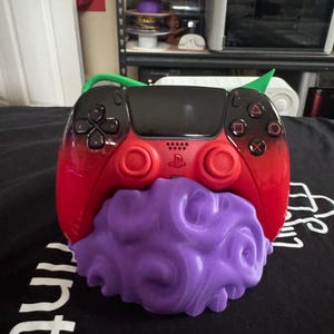 May include: A red and black gaming controller resting on a purple, textured, cloud-shaped stand. The controller features a black touchpad, black directional buttons, and red analog sticks. The stand has a swirling, organic surface design.