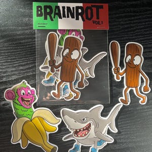 Könnte beinhalten: Eine Sammlung von Aufklebern mit Cartoon-Charakteren. Enthält einen Hai, einen Affen in einer Banane und eine Strichmännchenfigur mit einem Baseballschläger. Die Aufkleber sind mit "BRAINROT VOL. 1" beschriftet.