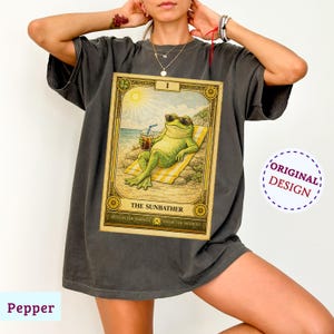 May include: Pepper-colored t-shirt featuring a tarot card design. The card depicts a frog in sunglasses relaxing on a beach towel, holding a drink. The card is framed with gold accents and the text "THE SUNBATHER". The shirt has a relaxed fit.