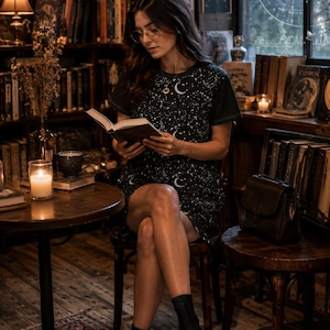 May include: A woman seated in a cozy library setting, reading a book. She wears a black dress adorned with a white celestial pattern. The room is filled with bookshelves, candles, and vintage decor, creating a warm and inviting atmosphere.