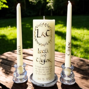 May include: A set of three off-white candles including one large pillar candle and two taper candles. The pillar candle features a gold laurel wreath with the letters L&C, the names Lucia and Taylor, a quote, and the date June 25, 2022. The taper candles are inscribed with the names Lucia and Taylor, and all three candles are held in clear glass candle holders.