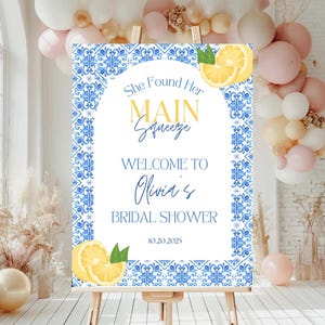 May include: A welcome sign for a bridal shower, featuring a blue and white tile pattern. The sign includes the text "She Found Her Main Squeeze, Welcome to Olivia's Bridal Shower" and lemon graphics. The date 10.20.2025 is also shown.