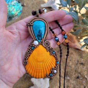 May include: A handmade pendant necklace with an orange seashell, decorated with turquoise and brown clay details. The pendant has a large turquoise stone and is suspended on a dark brown cord with orange and blue beads.