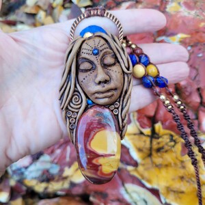 May include: A handcrafted pendant necklace featuring a sculpted face with closed eyes, adorned with a gemstone and beaded accents. The pendant is primarily brown with red, yellow, and blue details. The necklace is held by a brown cord.