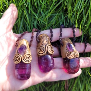 May include: Three pendant necklaces with polished purple gemstones. Each pendant has a gold-coloured metal cap with spiral patterns and a small amethyst crystal. The necklaces are suspended on brown cords. The background is green.