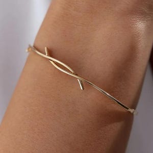 May include: A gold-colored bracelet with a minimalist design. The bracelet features a thin, curved band with a central decorative element composed of intersecting lines. The jewelry is worn on a wrist, showcasing its elegant simplicity.