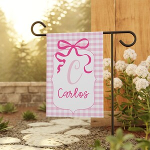May include: A pink and white gingham garden flag featuring a decorative bow and the letter "C" in light pink. The name "Carlos" is written in cursive below the letter. The flag is displayed on a black metal garden stake.