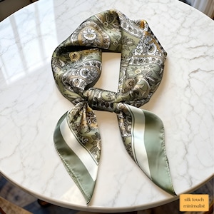 Peut inclure: Un foulard en soie vert avec un motif cachemire dans les tons beige et blanc. Le foulard est noué, avec les extrémités pendantes. Il présente une bordure rayée et le texte "silk touch minimalist".